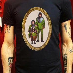 May include: A black short-sleeved t-shirt featuring a central oval graphic framed in gold. The print depicts a family of three anthropomorphic chimpanzees dressed in green and brown tunics, standing and sitting in a portrait style.