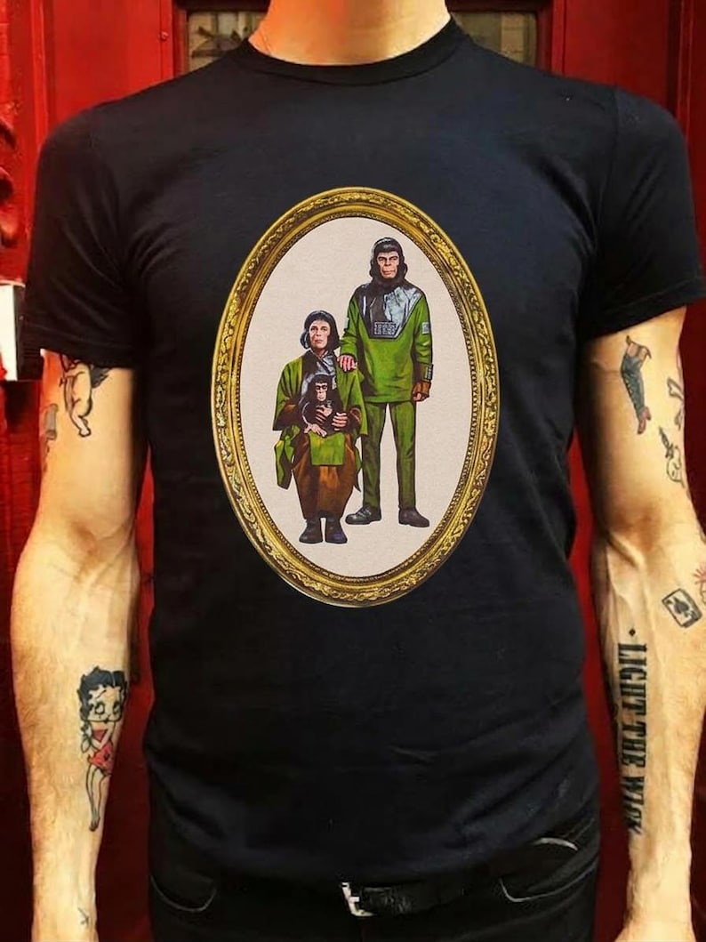 May include: A black short-sleeved t-shirt featuring a central oval graphic framed in gold. The print depicts a family of three anthropomorphic chimpanzees dressed in green and brown tunics, standing and sitting in a portrait style.