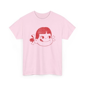 A flat lay of a light pink t-shirt with a red graphic of a girl's face with pigtails and a winking expression.