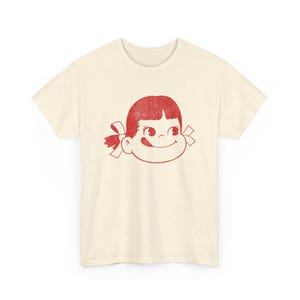 A flat lay of a cream-colored t-shirt with a red graphic of a girl's face with pigtails and a winking expression.