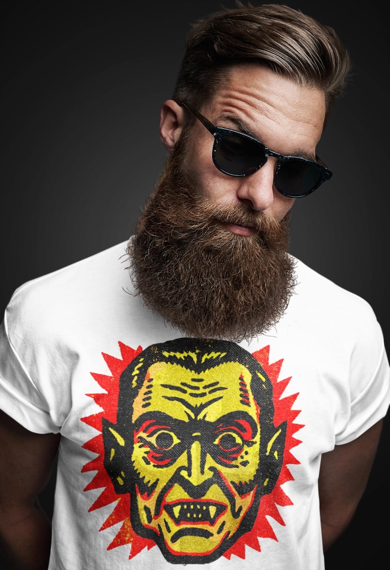May include: White t-shirt featuring a vintage-style illustration of a vampire's face in yellow and black, with a red burst background. The t-shirt is worn by a person with a long beard and sunglasses.