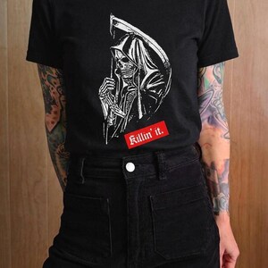 A person wearing a black t-shirt with a white grim reaper graphic and a red rectangular label with white text.
