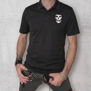 A man wearing a black polo shirt with a white skull logo on the left chest, paired with dark distressed jeans and a studded belt.