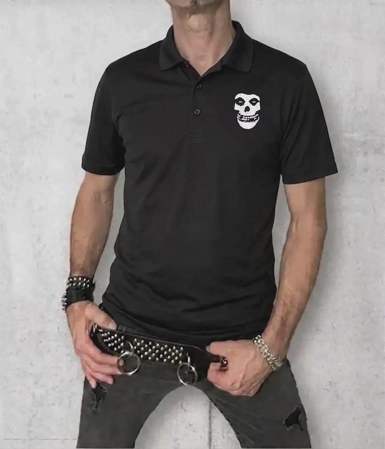 A man wearing a black polo shirt with a white skull logo on the left chest, paired with dark distressed jeans and a studded belt.