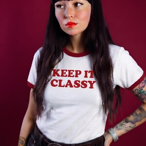 May include: A white ringer t-shirt with short sleeves and a red crew neckline. The text "KEEP IT CLASSY" is printed in red on the chest. The shirt is paired with dark pants and a brown leather belt featuring a large, round, metallic buckle.