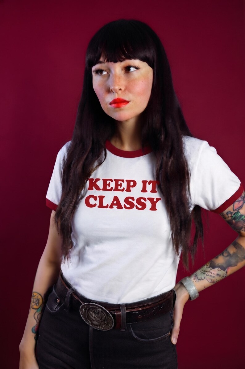 May include: A white ringer t-shirt with short sleeves and a red crew neckline. The text "KEEP IT CLASSY" is printed in red on the chest. The shirt is paired with dark pants and a brown leather belt featuring a large, round, metallic buckle.