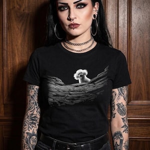 May include: A black short-sleeved t-shirt featuring a graphic print of a small white fluffy dog sitting on a textured, rocky, grey landscape. The shirt has a crew neckline and is made of a solid black fabric, providing a stark contrast to the central image.