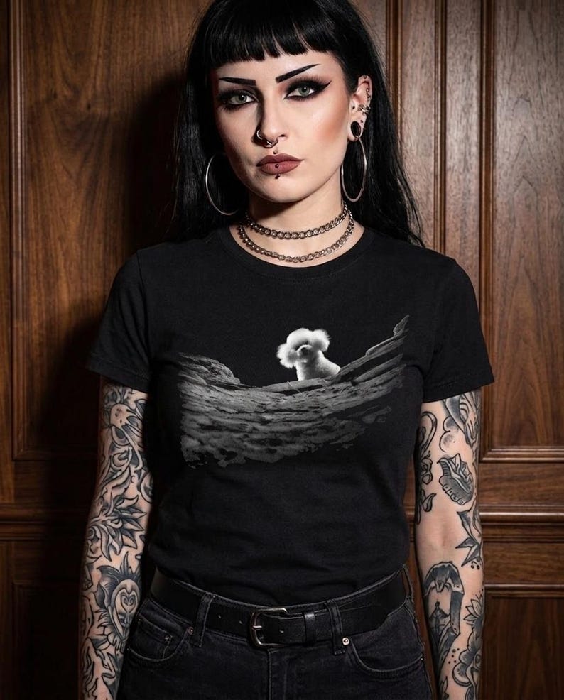 May include: A black short-sleeved t-shirt featuring a graphic print of a small white fluffy dog sitting on a textured, rocky, grey landscape. The shirt has a crew neckline and is made of a solid black fabric, providing a stark contrast to the central image.