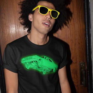 May include: A black short-sleeve t-shirt featuring a glowing neon green graphic of a vintage car on the chest. The shirt has a crew neckline and is worn with bright yellow plastic sunglasses with dark lenses, creating a high-contrast look.