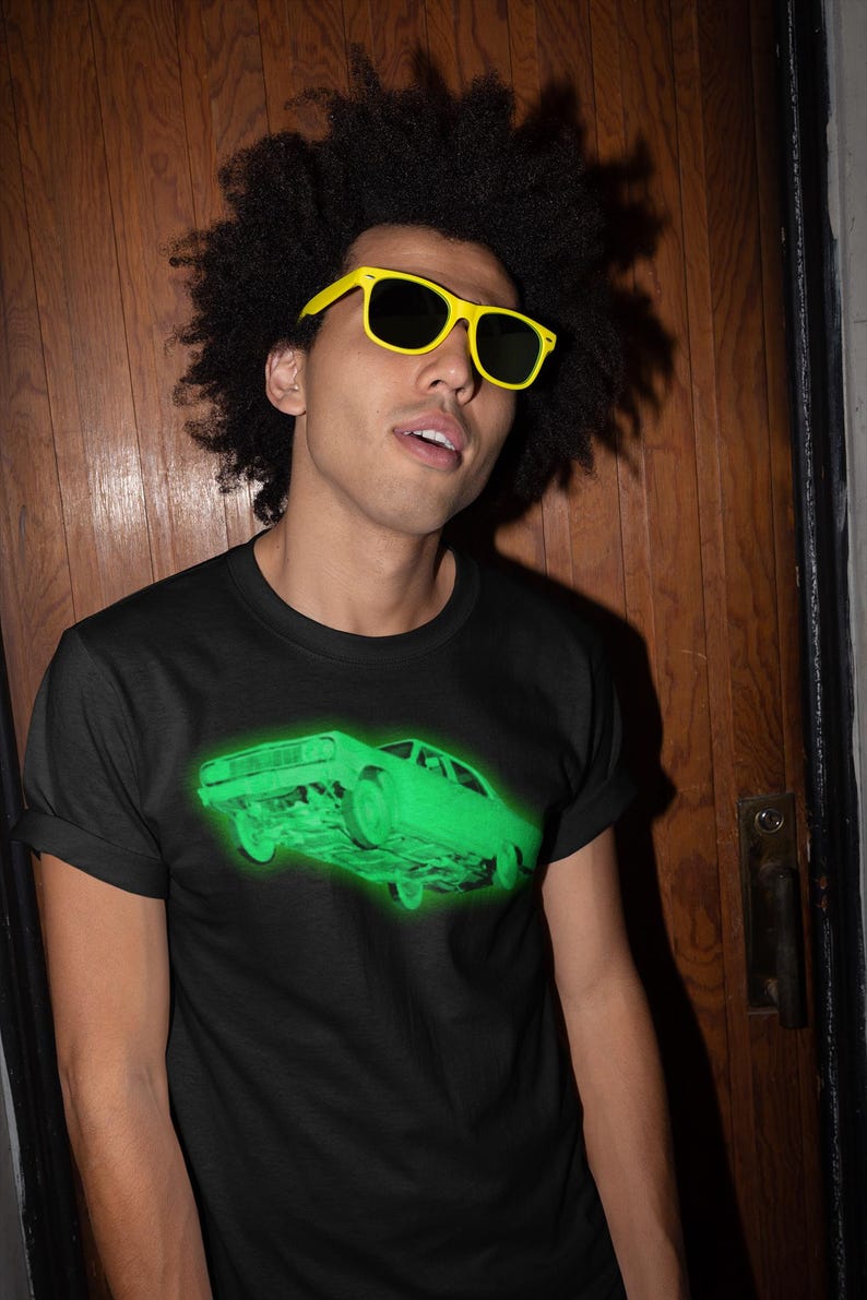 May include: A black short-sleeve t-shirt featuring a glowing neon green graphic of a vintage car on the chest. The shirt has a crew neckline and is worn with bright yellow plastic sunglasses with dark lenses, creating a high-contrast look.