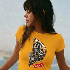 A person wearing a yellow fitted t-shirt with the grim reaper graphic and Killin it text, standing outdoors with a blurred background.