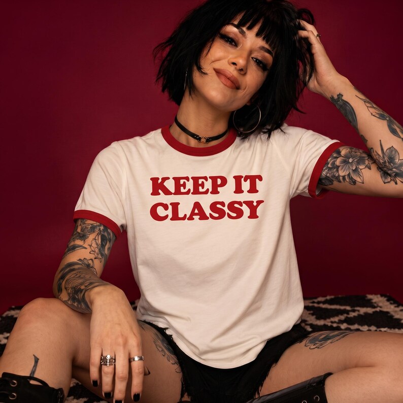 May include: A white ringer t-shirt with red trim on the collar and sleeves. The shirt features the red text "KEEP IT CLASSY" printed across the chest in a bold, sans-serif font. The garment has short sleeves and a crew neckline.