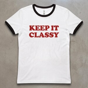 May include: A white ringer t-shirt with black trim on the collar and sleeve cuffs. The shirt features the text KEEP IT CLASSY printed in a bold, red, serif font across the center of the chest area, creating a high-contrast graphic design.