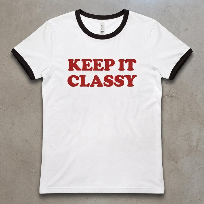 May include: A white ringer t-shirt with black trim on the collar and sleeve cuffs. The shirt features the text KEEP IT CLASSY printed in a bold, red, serif font across the center of the chest area, creating a high-contrast graphic design.