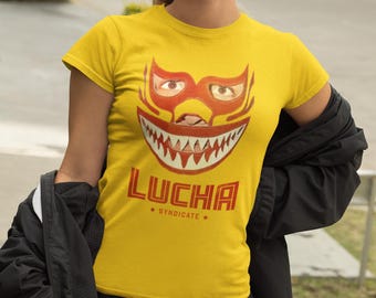 May include: A yellow t-shirt with a red and orange luchador mask design and the word "LUCHA" in red. The text "* SYNDICATE *" is below the word "LUCHA". The mask has sharp teeth.