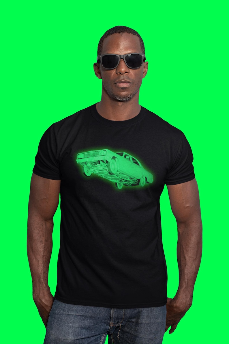 May include: A black short-sleeved t-shirt featuring a glowing neon green graphic of a vintage sedan car on the chest. The car illustration is rendered in a wireframe style, showing the front and side profile of the vehicle in a bright, luminous green hue.