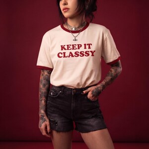 May include: A cream-colored ringer t-shirt with dark red trim on the collar and sleeves. The text KEEP IT CLASSSY is printed in dark red on the chest. The shirt is paired with black denim shorts featuring a distressed, frayed hemline.