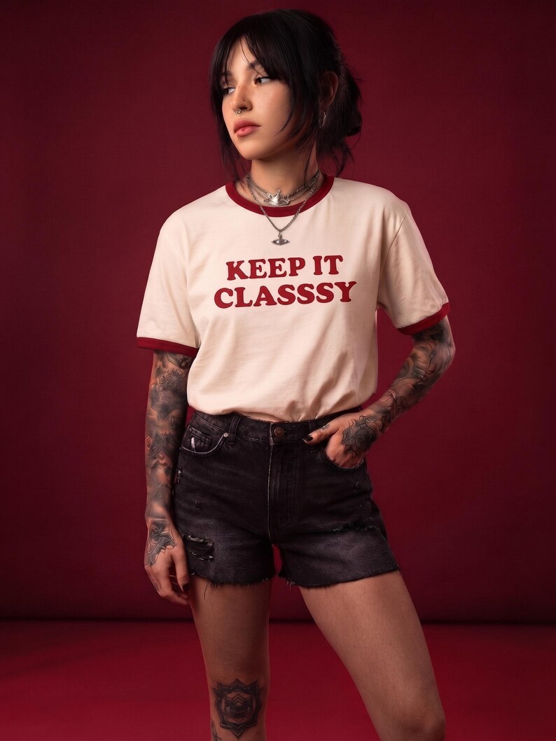 May include: A cream-colored ringer t-shirt with dark red trim on the collar and sleeves. The text KEEP IT CLASSSY is printed in dark red on the chest. The shirt is paired with black denim shorts featuring a distressed, frayed hemline.