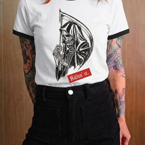A person with bright red hair wearing a white ringer t-shirt with black trim and a graphic of the grim reaper with a red Killin it caption.