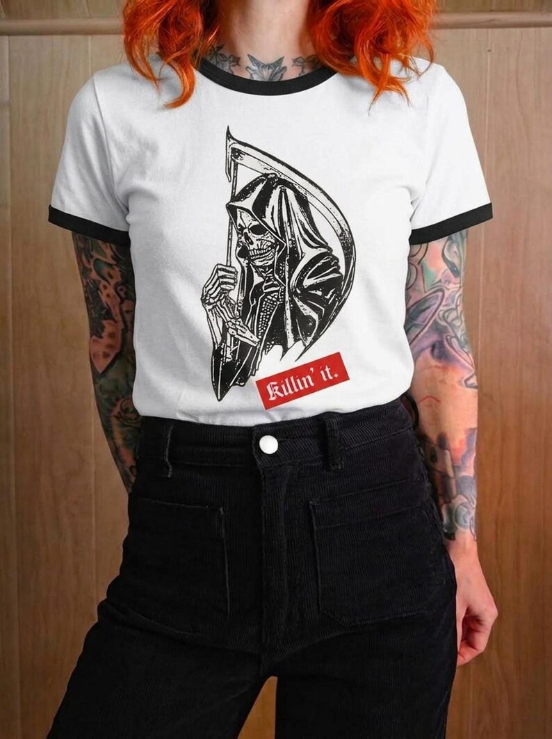 A person with bright red hair wearing a white ringer t-shirt with black trim and a graphic of the grim reaper with a red Killin it caption.