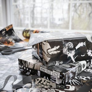 Two stacked gifts wrapped in black and white Misfits-themed paper, with loose paper and scissors on a table by a window.