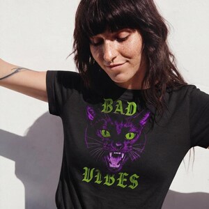 A person with dark hair and freckles looking down while wearing the black graphic t-shirt against a light wall.