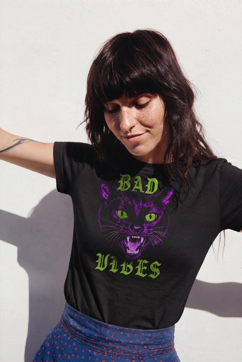 A person with dark hair and freckles looking down while wearing the black graphic t-shirt against a light wall.