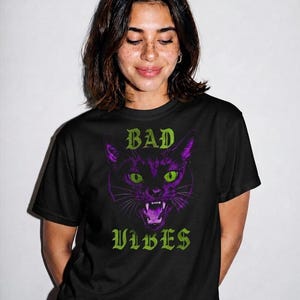 A smiling person with freckles and shoulder-length dark hair wearing the black t-shirt with the purple cat graphic.