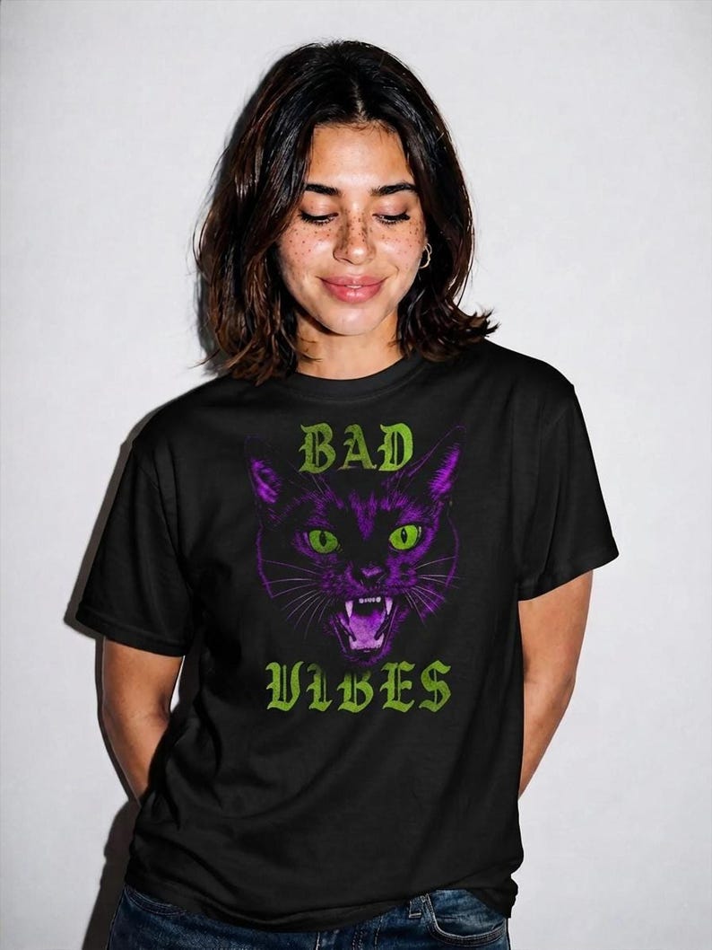 A smiling person with freckles and shoulder-length dark hair wearing the black t-shirt with the purple cat graphic.