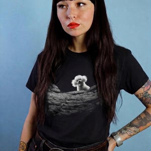 May include: A black short-sleeved t-shirt featuring a graphic print of a white fluffy dog sitting on a textured, cratered lunar surface. The design is centred on the chest area of the shirt, contrasting against the solid black fabric background.