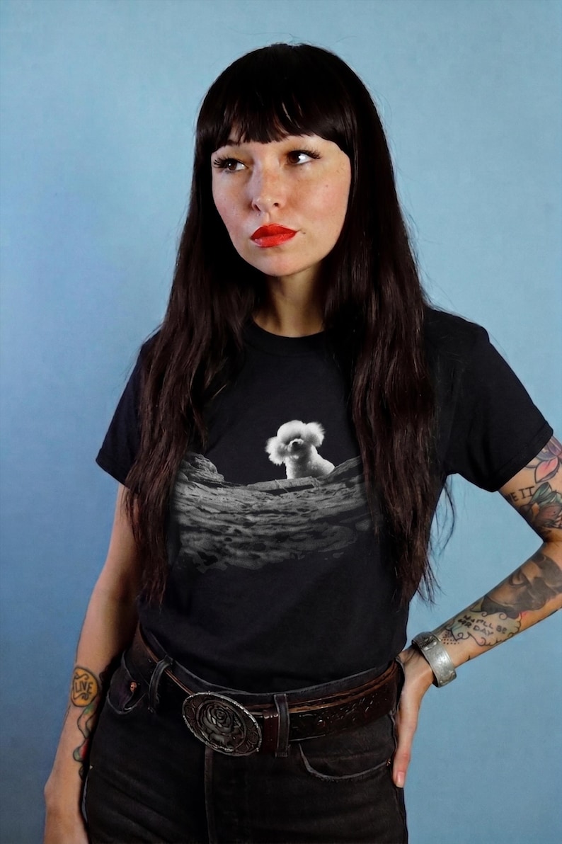 May include: A black short-sleeved t-shirt featuring a graphic print of a white fluffy dog sitting on a textured, cratered lunar surface. The design is centred on the chest area of the shirt, contrasting against the solid black fabric background.