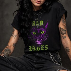 May include: A black short-sleeved t-shirt featuring a graphic of a purple cat with bright green eyes and an open mouth. The text "BAD" appears above the cat and "VIBES" appears below in a green gothic font. The shirt has a crew neckline.