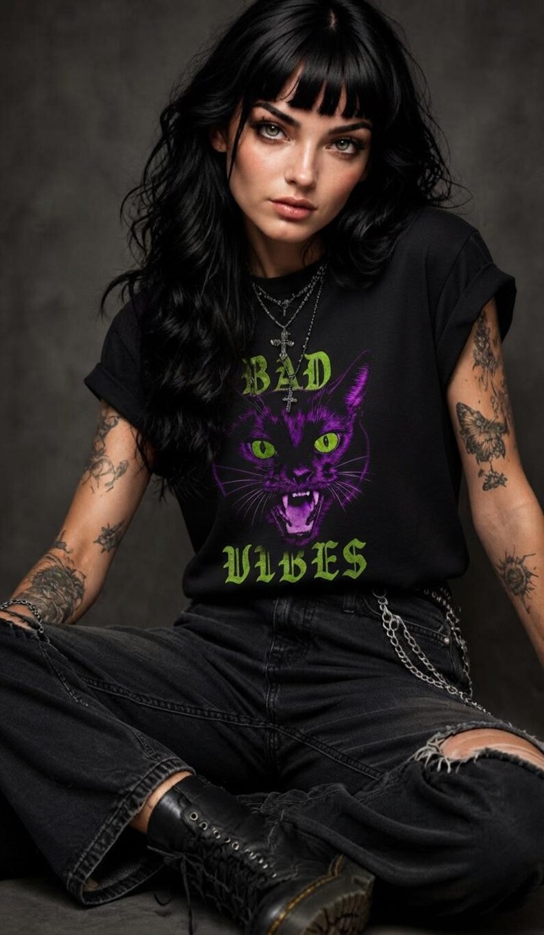 May include: A black short-sleeved t-shirt featuring a graphic of a purple cat with bright green eyes and an open mouth. The text "BAD" appears above the cat and "VIBES" appears below in a green gothic font. The shirt has a crew neckline.