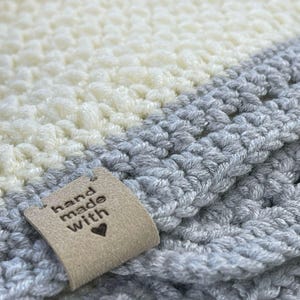 May include: A close-up of a gray and white crocheted blanket with a brown leather tag that says "hand made with" and a small heart.