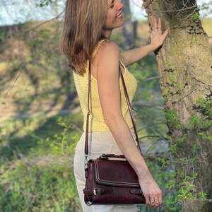 Peut inclure: Une femme portant un d&eacute;bardeur en crochet jaune et un pantalon blanc se tient &agrave; c&ocirc;t&eacute; d'un arbre. Elle tient un sac &agrave; main en cuir marron avec une bandouli&egrave;re.