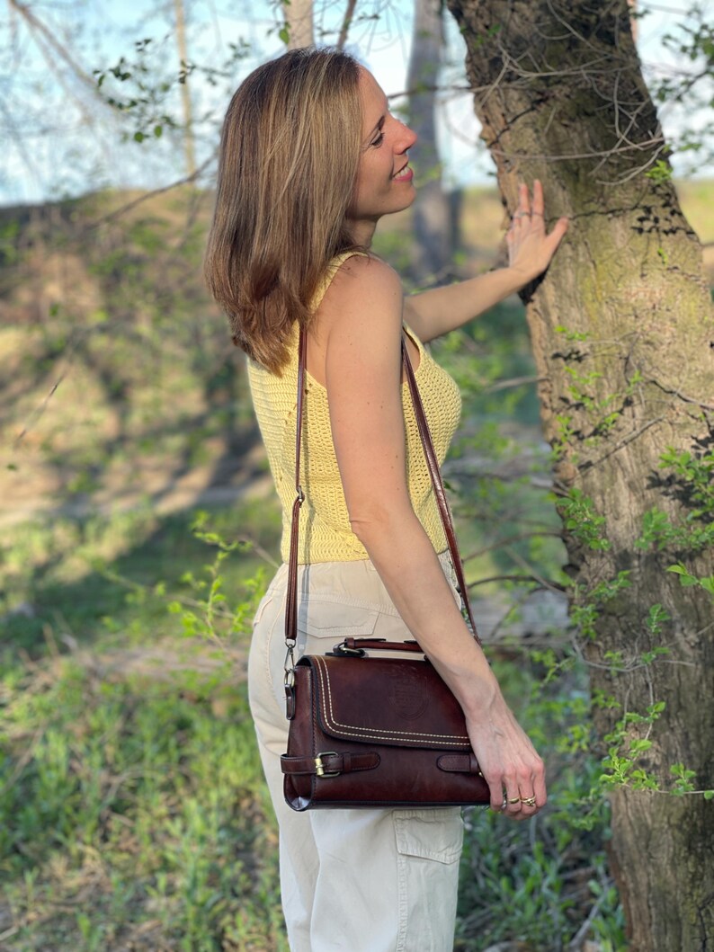 Peut inclure: Une femme portant un d&eacute;bardeur en crochet jaune et un pantalon blanc se tient &agrave; c&ocirc;t&eacute; d'un arbre. Elle tient un sac &agrave; main en cuir marron avec une bandouli&egrave;re.