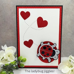 May include: A white card with a red border and red glitter hearts. A ladybug with black spots is on the card with the text "The ladybug jiggles!"