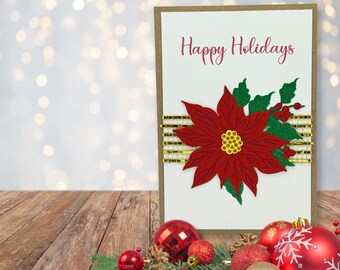 May include: A rectangular greeting card with a kraft paper border and white center featuring the red text Happy Holidays. A large red poinsettia with green leaves and a yellow center sits over a horizontal strip of gold metallic mesh ribbon.
