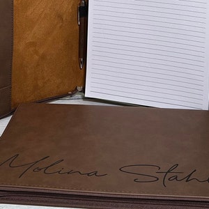 May include: Brown leather zippered portfolio with a pen holder and lined notepad. The portfolio is personalized with the name "Molina Stahl" in a cursive script.