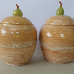 Könnte beinhalten: Zwei Keramik-Behälter mit birnenförmigen Deckeln. Die Behälter sind hellbraun mit weißen Streifen und einer glänzenden Oberfläche. Die Deckel sind hellgrün.