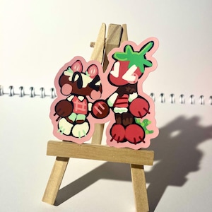 Puede incluir: Dos pegatinas de dibujos animados en un pequeño caballete de madera. Una pegatina presenta un personaje marrón con un lazo rosa y un atuendo blanco y verde. La otra pegatina es un personaje de fresa con una parte superior verde y un cuerpo rojo. Ambas pegatinas se dan la mano.