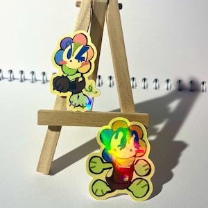 Puede incluir: Dos pegatinas holográficas con un personaje de dibujos animados con cabeza en forma de flor y cara sonriente. Las pegatinas se exhiben en un pequeño caballete de madera. El personaje tiene un diseño colorido con un contorno amarillo.