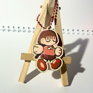 以下が含まれることがあります： ピンクのシャツと茶色のスカートを着た、目を閉じた漫画のキャラクターが特徴の木製キーホルダー。 キーホルダーは赤いビーズチェーンに取り付けられ、小さな木製イーゼルに展示されています。