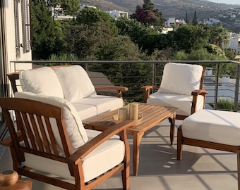 May include: Outdoor wooden furniture set with white cushions. The set includes a loveseat, two armchairs, a small coffee table, and an ottoman. The furniture is on a balcony with a view of trees and buildings.