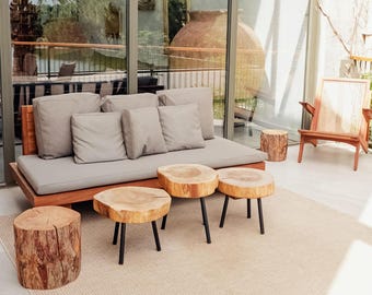May include: Outdoor patio furniture set featuring a wooden sofa with gray cushions, wooden side tables, and tree stump stools. A wooden chair with a woven back and a neutral rug complete the scene. The setting is bright and airy.