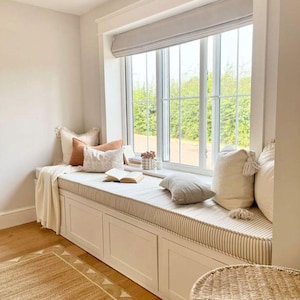 May include: A window seat with a striped cushion, several pillows, and a cream-colored blanket. The built-in storage bench is painted white. An open book and a small vase of flowers create a relaxing space. The window has a light-colored blind.