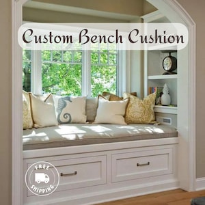 May include: A white window seat with a beige cushion and pillows. The seat has two drawers and is built into a bay window. The text "Custom Bench Cushion" is displayed above the window.