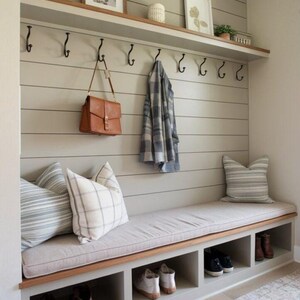 May include: A built-in entryway bench with a gray cushion, storage cubbies, and a wooden trim. Above, a shelf with decor and black hooks for hanging items. A brown leather bag and a plaid scarf hang from the hooks.