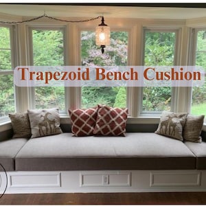 Op de afbeelding: Een grijze trapeziumvormige bankkussen met een witte basis en drie laden. Het kussen is bekleed met een lichtgrijze stof en heeft drie kussens erop. De kussens zijn lichtbruin, rood-wit geruit en lichtbruin met een giraffenprint. De bank staat voor een erkerraam met uitzicht op bomen. De tekst "Trapezoid Bench Cushion" is geschreven in rode letters op een witte achtergrond.