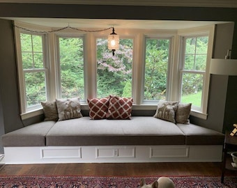 May include: A built-in window seat with a white base featuring rectangular paneling, topped with a long, light gray cushion. The seat is decorated with two beige pillows featuring giraffe illustrations and two rust-colored pillows with a white lattice pattern.